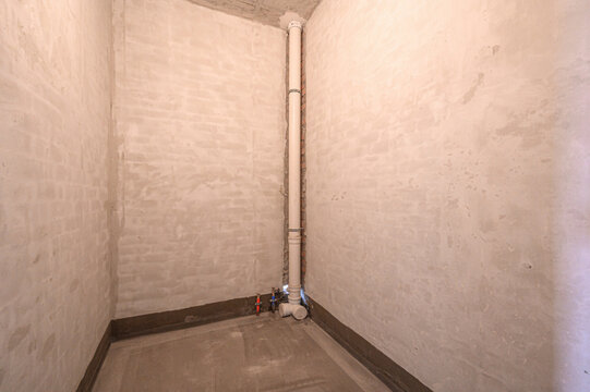  Unfinished bathroom with plastered walls, concrete floor, exposed plumbing pipe, water supply valves, corner layout, hydroisolation layer visible, ready for tiling.