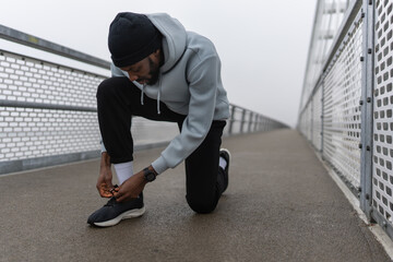 Fototapeta premium Black male runner kneeling to tie his shoe on a foggy urban bridge, wearing hoodie and beanie, outdoor workout preparation, focus, discipline and determination