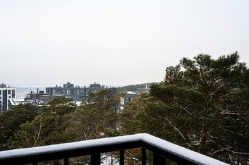 Fototapeta premium Balcony view of modern residential buildings surrounded by pine trees, light snow on railing, overcast sky, serene suburban atmosphere.