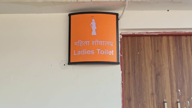 Tiltup shot of an orange ladies toilet signboard with Hindi and English text mounted on a station wall, clearly indicating women restroom facility inside premises