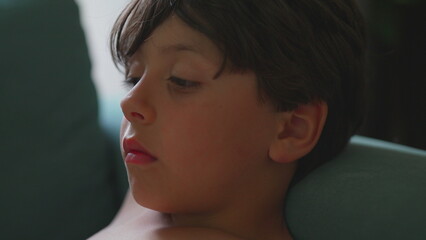Young boy with serious expression resting on armchair indoors, natural light from window highlighting quiet mood and focused gaze © Marco