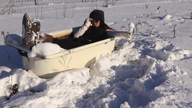 Beautiful girl resting in an old bathtub in the open air in the snow, playfully filling a glass with snow. The concept of contrast between winter cold and relaxation.