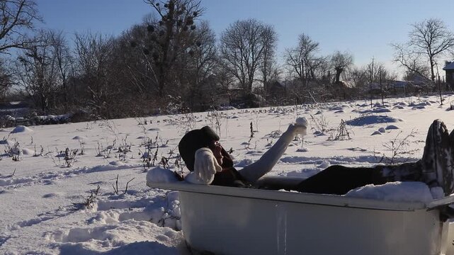 Beautiful girl resting in an old bathtub in the open air in the snow, playfully filling a glass with snow. The concept of contrast between winter cold and relaxation.