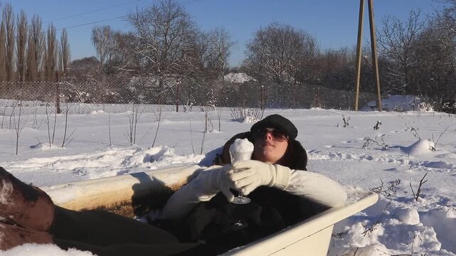 Creative winter lifestyle shot: a woman lies in an open-air bathtub amidst the snow, holding a glass of snow in the form of a cocktail