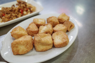 A close-up shot of several deep-fried crispy tofu cubes served on an oval white ceramic plate. The tofu has a golden brown texture and is presented as a healthy vegetarian appetizer in an indoor.
