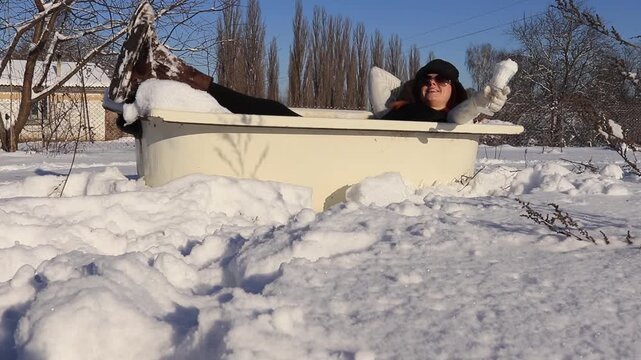 A woman with red hair in winter clothes sits in a snowy bathtub in the yard and holds a shovel, a humorous scene on a frosty sunny day.