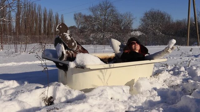 Beautiful girl resting in an old bathtub in the open air in the snow, playfully filling a glass with snow. The concept of contrast between winter cold and relaxation.