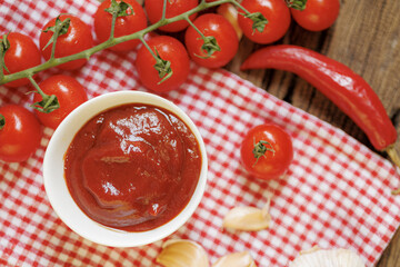 Bowl of rich tomato sauce sits on red and white checkered cloth, surrounded by fresh cherry tomatoes, red chili pepper, and garlic cloves, creating rustic and appetizing scene