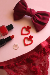 Red lace bralette, hairbow, heart shaped earrings, golden ring, red lipstick and nail polish on pink background. Coquettish accessories for Valentine's day. Selective focus.