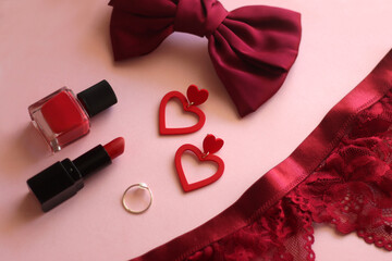 Red lace bralette, hairbow, heart shaped earrings, golden ring, red lipstick and nail polish on pink background. Coquettish accessories for Valentine's day. Selective focus.