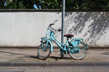 A light blue city bicycle leans against a pole with white wall in the background