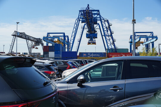 Used cars parked near harbor cranes with stacked containers forming an industrial backdrop