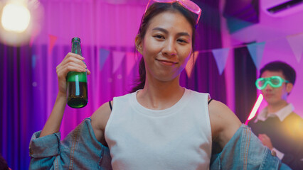 Diverse multi ethnic young adult Asian lady wear futuristic neon glowing glasses smile and drink alcohol beer fun enjoy hangout party in living room night indoor home. Disco lights illuminating house.
