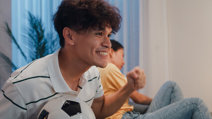 Asian new marriage couple sit on sofa husband excited cheering watching football sport game on television in living room at home. Young woman getting bored her partner watch online soccer on TV.