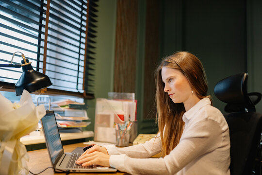 Woman works on laptop at wooden desk in home office during day, concentrating on the screen