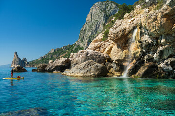 Man paddleboarding on crystal-clear turquoise sea along the rocky coast of Baunei, Sardinia, with a small waterfall cascading into the Mediterranean Sea on a sunny summer day