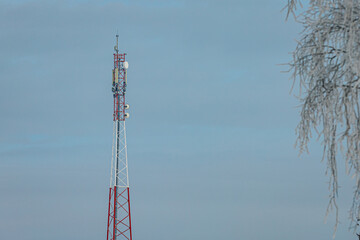 Obraz premium Telecommunications tower with red and white structure against a cloudy sky, surrounded by frost-covered branches in a winter landscape