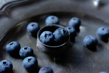 Moody dark-tone close-up of fresh blueberries arranged in a small vintage metal bowl and scattered...
