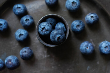 Moody dark-tone close-up of fresh blueberries arranged in a small vintage metal bowl and scattered...