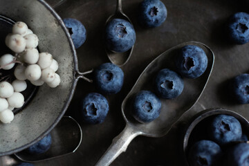 Moody dark-tone close-up of fresh blueberries arranged in a small vintage metal bowl and scattered...