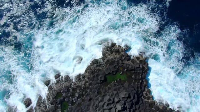 Overhead drone video: powerful turquoise, sapphire ocean waves crash dark basaltic rocks, exploding white foam under brilliant high-key daylight. Majestic nature power