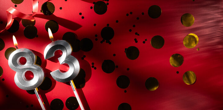 Concept of celebrating birthday and anniversary. Number 83 eighty-three silver celebration birthday candle on festive red background with large golden confetti. Eighty-three years birthday.
