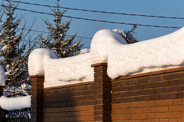 Thick snowdrifts cap a brick fence, creating a winter wonderland scene. The sun shines brightly on...