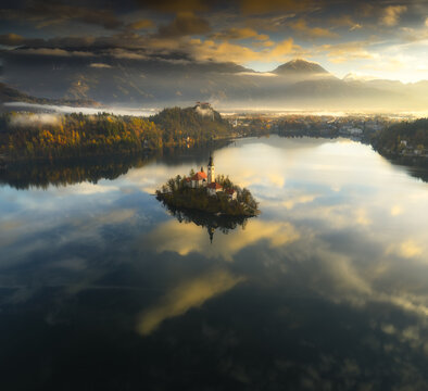 Aerial view of the island church standing peacefully in the middle of the lake under a sky of orange and gray, Spodnja Lua, kofja Loka, Slovenia.