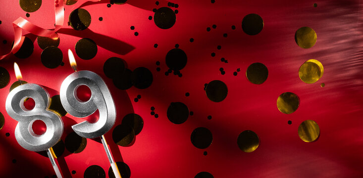 Concept of celebrating birthday and anniversary. Number 89 eighty-nine silver celebration birthday candle on festive red background with large golden confetti. Eighty-nine years birthday.