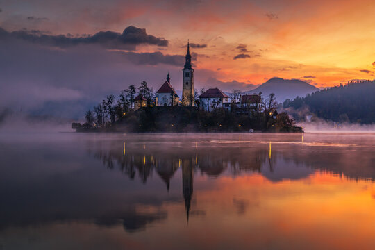 Aerial view of island church reflected in tranquil waters under a vibrant sky painted with hues of orange and purple, Spodnja Lua, kofja Loka, Slovenia.