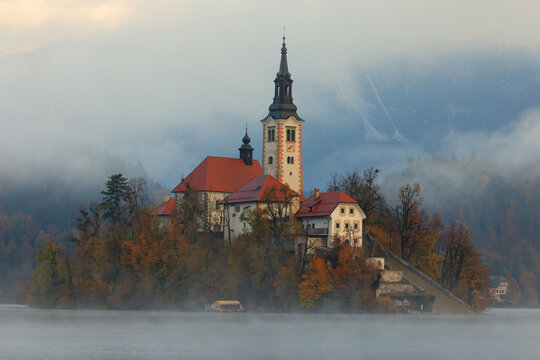 Aerial view of the iconic church standing proudly on a small island amidst the tranquil lake waters, cloaked in a mystical veil of fog, Spodnja Lua, kofja Loka, Slovenia.