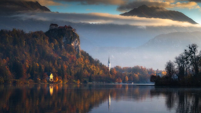 Aerial view of Lake Bled reflects the castle perched atop a cliff, autumn colors painting the landscape under a misty sky, Spodnja Lua, kofja Loka, Slovenia.