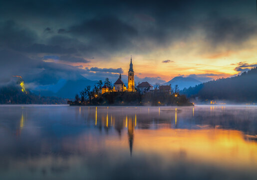 Aerial view of Island with the Church of the Assumption mirroring in the tranquil waters, under a vibrant sky at dusk, Spodnja Lua, Slovenia.