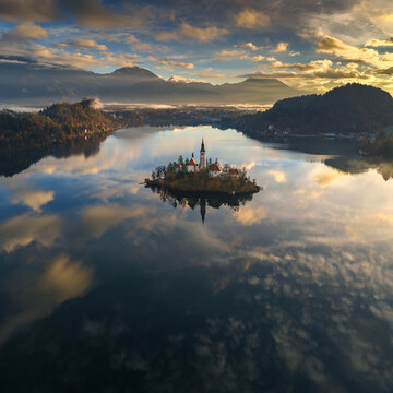 Aerial view of an island church mirrored in the tranquil lake's surface, framed by distant mountains and a dramatic sky, Spodnja Lua, kofja Loka, Slovenia.