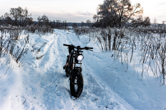 Robust electric fat bike, headlight glowing, navigates fresh powder on a serene winter trail. A cool ride for snowy adventures