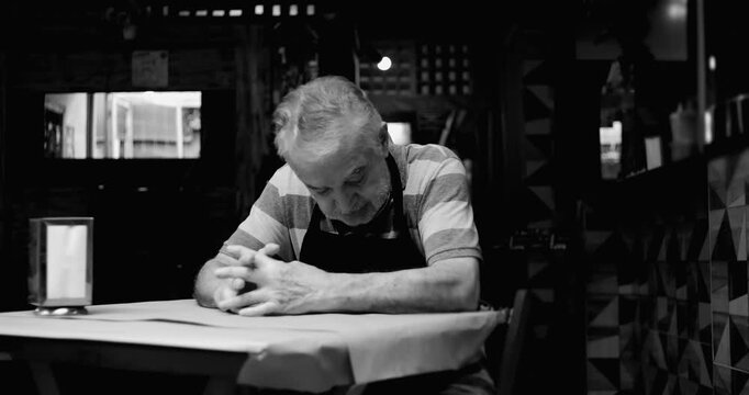 Older caf&eacute; owner sits in silence at table with hands clasped conveying uncertainty fatigue and bleak acceptance during difficult period for small business in black and white