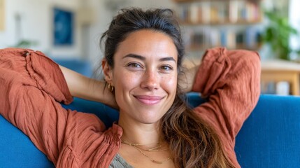 Young woman leans back on blue sofa with hands behind her head, smiling in bright living room with daylight and bookshelves in background