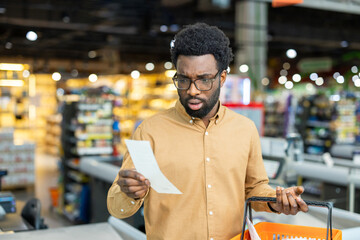Naklejka premium Confused man looking at a long grocery store receipt, expressing shock and worry about rising prices and inflation, standing with a shopping basket at the checkout counter