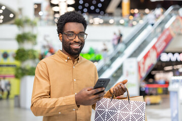 Man smiling at camera while carrying shopping bags and checking smartphone with a blurred shopping...