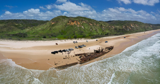 Aerial view of the Maheno shipwreck resting on golden sands, juxtaposed against the azure sea and green dunes, K'Gari Fraser Island, Queensland, Australia.