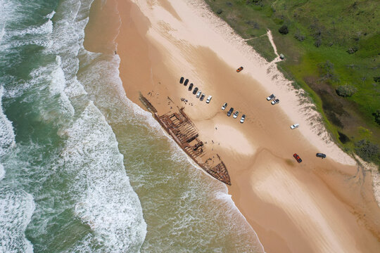 Aerial view of Maheno rusted shipwreck resting on golden sands, met by azure waves and backed by lush greenery, K'Gari Fraser Island, Queensland, Australia.