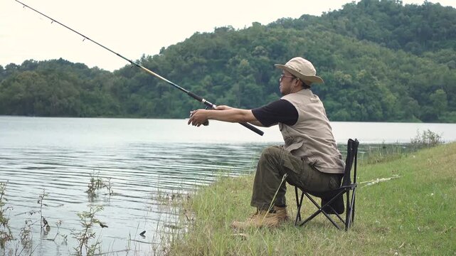 A survivalist sitting on a collapsible chair at the lake&rsquo;s edge, casting a fishing line with a spinning reel. Outdoor survival, fishing activity, and peaceful nature lifestyle