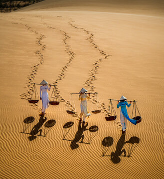 Aerial view of three Vietnamese women in traditional ao dai costume, carrying straw baskets whilst transversing a sand dune. Their shadows cast long. Mui Ne, Phan Thiet, Vietnam.