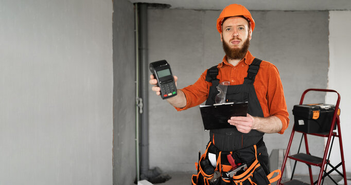 Portrait of bearded builder in hardhat holding POS terminal for contactless payment.