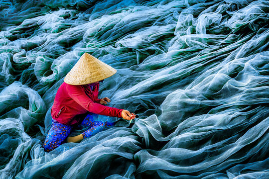 Aerial view of a woman in traditional conical hat mending fishing nets, a sea of turquoise textures under the bright sun, Phan Rang, Thap Cham, Vietnam.