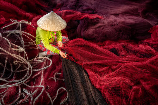 Aerial view of a woman with a conical hat repairing fishing nets, amidst a sea of deep reds and blacks, Phan Rang, Thap Cham, Vietnam.