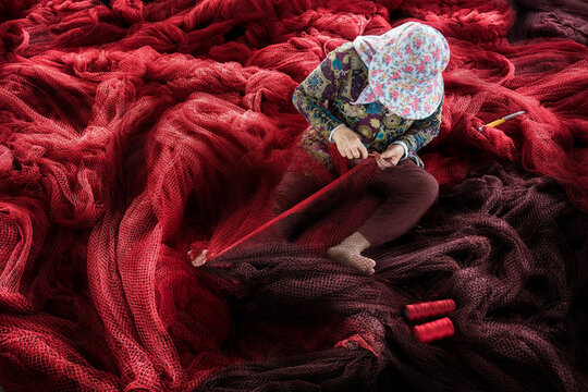 Aerial view of a woman meticulously mending vibrant red fishing nets, the deep crimson hues contrasting with her floral patterned attire, Phan Rang, Thap Cham, Vietnam.