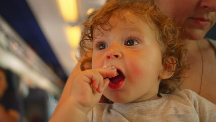 Curly-haired toddler bites into chocolate bar while looking up, seated on mother&rsquo;s lap inside moving train with cabin lights glowing