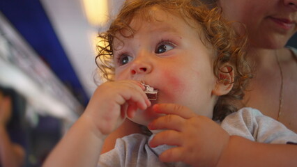 Curly-haired toddler bites into chocolate bar while looking up, seated on mother&rsquo;s lap inside moving train with cabin lights glowing