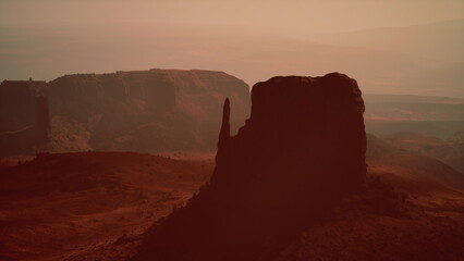 Fototapeta premium The sun sets behind impressive rock formations in a vast desert landscape, creating an enchanting atmosphere with hues of orange and brown. Shadows lengthen as night approaches.